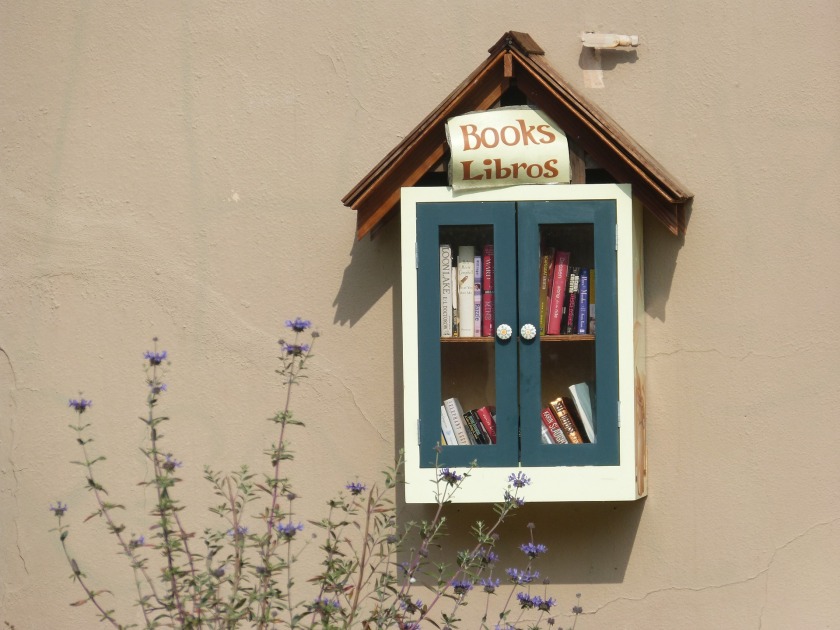 A little library on the side of a wall with flowers in front of it.